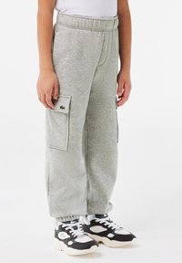 Pantalon de survêtement gris avec taille à cordon, poches latérales et un petit logo vert. Associé à des baskets de sport noir et blanc.