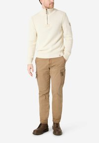 Beiger Strickpullover mit hohem Kragen und halbem Reißverschluss, kombiniert mit hellbraunen Cargohosen mit seitlichen Reißverschlusstaschen und dunkelbraunen Stiefeln.