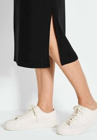 Robe noire mi-longue avec fentes sur les côtés, assortie à des baskets blanches à lacets. La robe présente des coutures impeccables et une texture lisse.