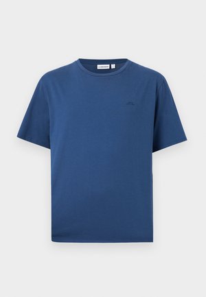 Noor mees seisab, kandes sinist lühikese varrukaga T-särki, musti lühikesi pükse, valgeid sokke ja beeže tossusid ühtlasel heledal taustal.