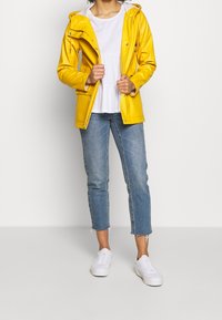 Žlutá waterproof bunda s kapucí, metalické akcenty, nošená přes bílý peplum top a světlé modré roztrhané džíny. Bílé tenisky doplňují outfit.