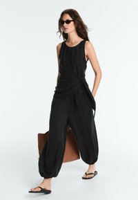 Femme portant une combinaison noire sans manches, des lunettes de soleil noires, des tongs noires, portant un grand sac fourre-tout marron, marchant devant un fond blanc.
