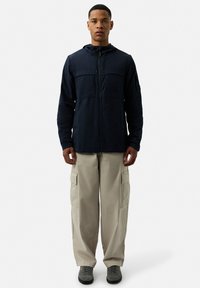 Navy hooded jacket with front zip, two chest pockets, and lightweight fabric, paired with beige cargo pants and gray shoes.