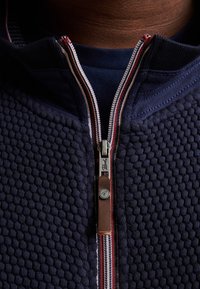 Marinblå zip-up jacka i texturerat stickat tyg, med en kontrasterande dragkedja och ett dragområde i brunt läder samt röda och vita randiga accenter.