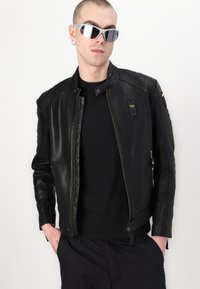 Giacca da moto in pelle nera con colletto alto, chiusura con zip, tasche laterali e dettagli a coste sulle spalle. Accompagnata da occhiali da sole argentati.