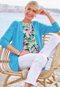 Türkiser Cardigan über einem blumengemusterten Oberteil, kombiniert mit weißen Hosen. Sitzt in einem geflochtenen Stuhl, der eine glatte Textur und einen lässigen Stil aufweist.