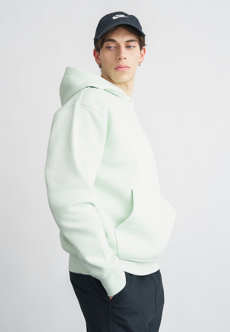 Μέντα πράσινο φούτερ με κουκούλα και τσέπη μπροστά, χαλαρή γραμμή και ριμπ μανσέτες. Φορεμένο με μαύρο καπέλο με λευκό λογότυπο.