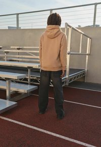 Beige hættetrøje med snøre, kombineret med mørke denimbukser, stående på en rød løbebane nær aluminiumsrækker, vendt væk.