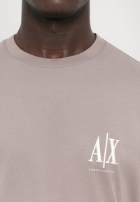 Világos bézs színű, rövid ujjú póló, puha anyagból készült, fehér hímzett „A/X” és „ARMANI EXCHANGE” logóval a mellkasán.