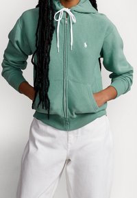 Felpa verde con zip realizzata in tessuto morbido, presenta un logo bianco sul petto, cappuccio con cordino regolabile e polsini e orlo a costine.