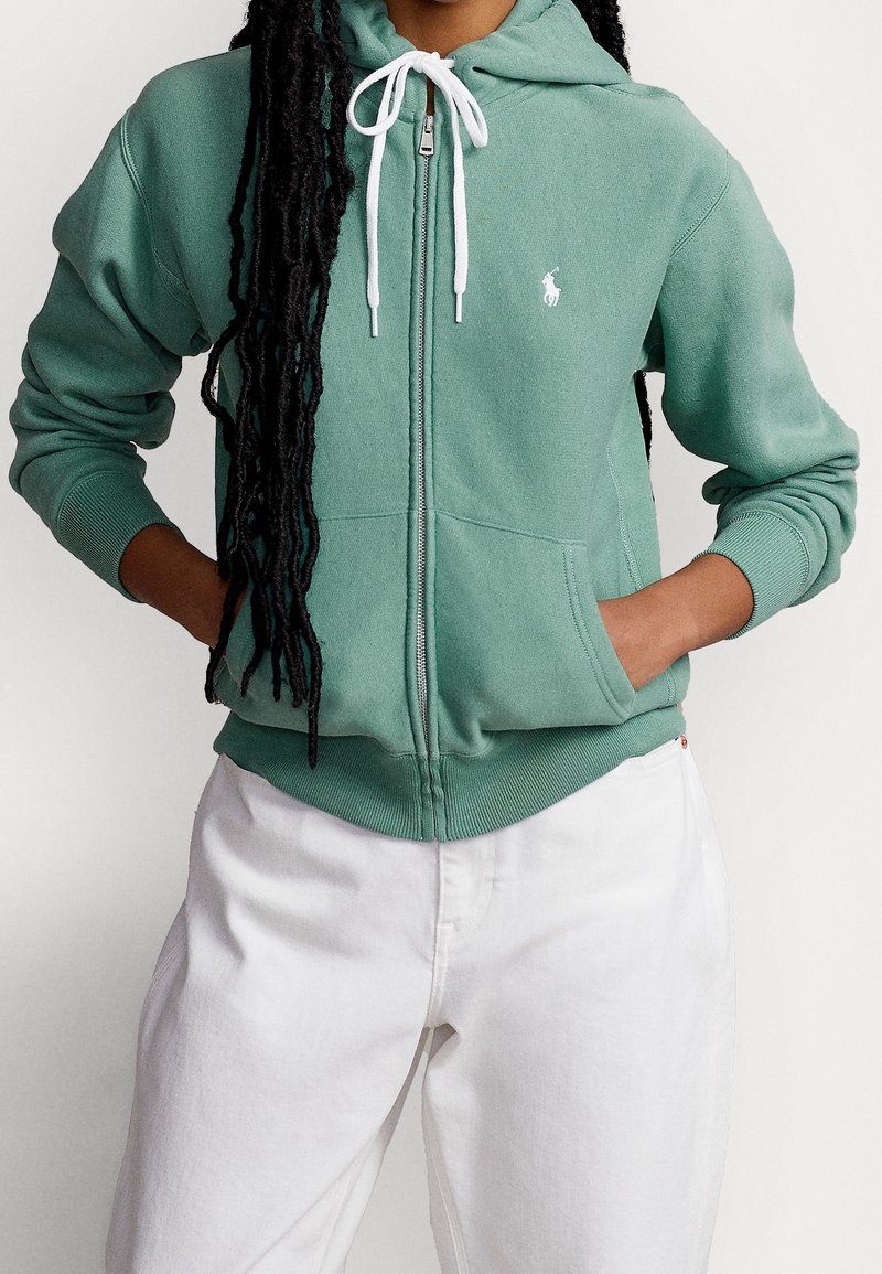 Felpa verde con zip realizzata in tessuto morbido, presenta un logo bianco sul petto, cappuccio con cordino regolabile e polsini e orlo a costine.