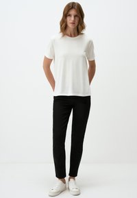 Witte T-shirt met korte mouwen en een ronde halslijn, gecombineerd met zwarte slim-fit broek en witte sneakers, weergegeven tegen een effen achtergrond.