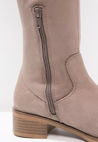 Gros plan d'une bottine en suède taupe avec une fermeture éclair latérale et un talon en bois empilé sur fond blanc.