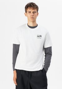 White cotton T-shirt with short sleeves layered over a long-sleeve gray shirt. Features a small black graphic logo on the chest. Black pants.