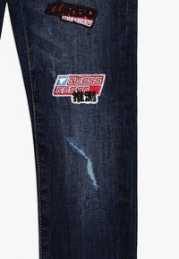 Tmavě modré džínové kalhoty s obnošenými záplatami. Mají vyšívané logo v červené, bílé a černé barvě, které přidává jedinečné vizuální prvky.