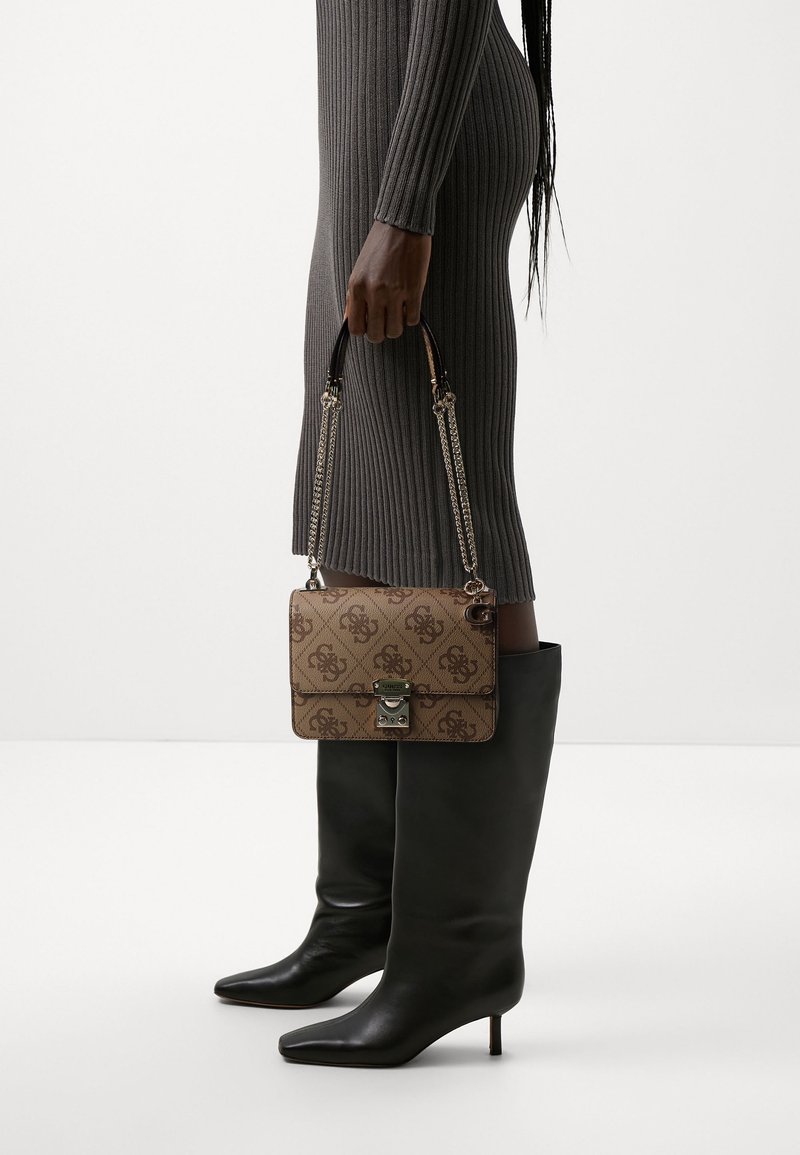 Personne portant une robe grise côtelée et des bottes noires à talons tient à ses côtés un sac à main beige à motifs avec une bandoulière chaîne.