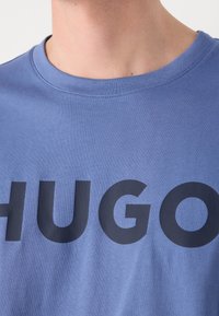 Gros plan sur une personne portant un T-shirt bleu avec de grandes lettres noires formant le mot "HUGO" sur la poitrine.