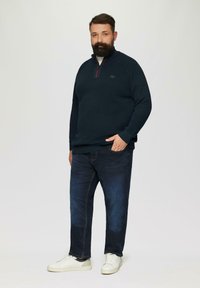 Marineblauwe ritstrui met een textuurgebreide stof, voorzien van een rode rits. Gecombineerd met donkere spijkerbroek en witte sneakers.