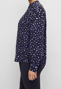 Langärmlige Bluse aus dunkelblauem Stoff mit lila und weißen abstrakten Mustern. Sie hat einen runden Halsausschnitt und Knopfdetails an den Ärmeln.