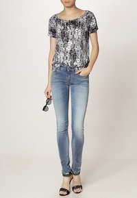 Top de manga corta con estampado en blanco y negro combinado con jeans skinny azul claro y sandalias de tiras negras; sosteniendo gafas de sol en una mano.