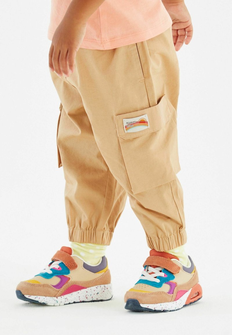 Enfant portant un pantalon cargo beige avec des bords élastiques et des baskets colorées, debout sur un fond blanc.