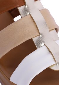 Sandalia con tiras que presenta cuero beige, marrón y blanco. Acento metálico dorado en las correas. Textura suave con punta redondeada.