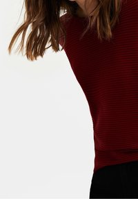 Haut en maille côtelée bordeaux avec un col rond et des manches courtes, coupe décontractée et texture lisse, partiellement rentré dans un pantalon foncé.