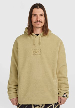 Man met schouderlang bruin haar, die een beige fleece-hoodie draagt met voorzakken en een klein geborduurd logo op de borst en mouw.