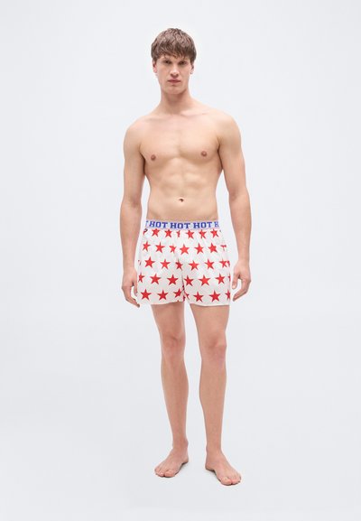 Κόκκινα και λευκά μποξέρ με μοτίβο αστέρια και μπλε μέση. Ελαφρύ ύφασμα, άνετη εφαρμογή. Φορεμένα από έναν αρσενικό μοντέλο που στέκεται ξυπόλυτος.