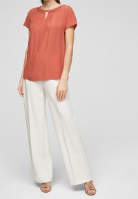 Blusa color ruggine con maniche corte, apertura a chiave e colletto ricamato. Abbinata a pantaloni bianchi a gamba larga e sandali beige con cinturini.