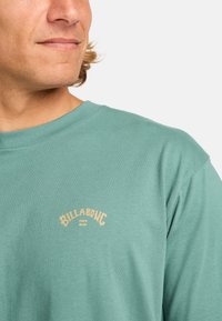 Camisa de manga corta verde menta con un tejido texturizado y un logotipo "Billabong" bordado en beige en el pecho.
