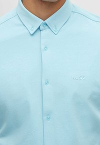 Hellblaues Hemd aus glattem Stoff mit spitzem Kragen, Markenlogo und klaren Kunststoffknöpfen.