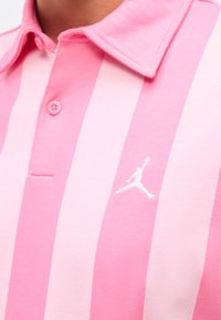 Gros plan sur un polo rayé rose et rose clair avec col, bouton et petit logo Jumpman blanc sur la poitrine.