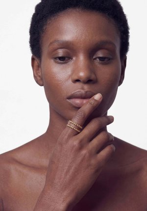 Femme aux cheveux courts et bouclés touchant ses lèvres, portant deux bagues dorées à perles sur son index, sur un fond blanc uni.