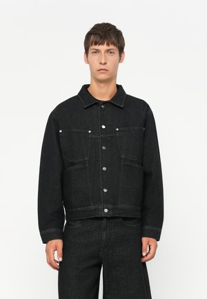 Young man wearing black denim jacket with button closure and matching black denim pants against a plain white background.