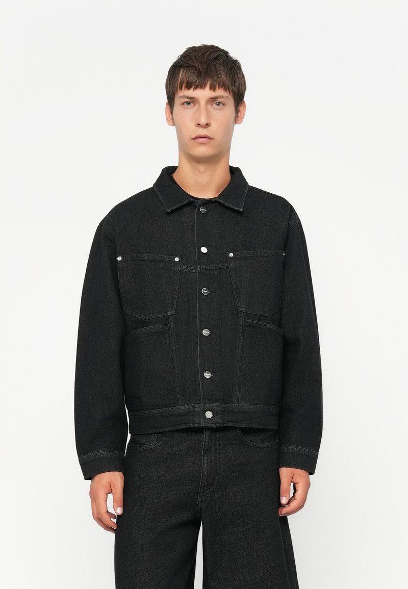 Jonge man in een zwarte denimjack met knoopsluiting en bijpassende zwarte denimbroek tegen een effen witte achtergrond.