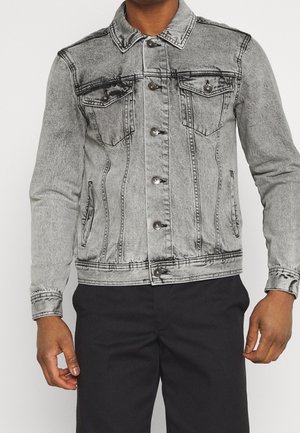 Mężczyzna w szarej acid-washowej kurtce jeansowej zapinanej na guziki i czarnych spodniach na jednolitym tle.