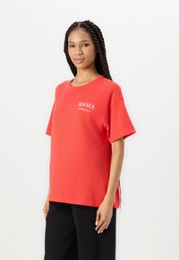 Camiseta de algodón roja con mangas cortas y cuello redondo, con el texto blanco "MAMA" en el pecho. Combinada con pantalones negros de canalé.