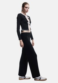 Svart ribbad crop top med vita detaljer, knuten framtill; svarta vida byxor och vita sneakers med svarta ränder och bruna sulor.