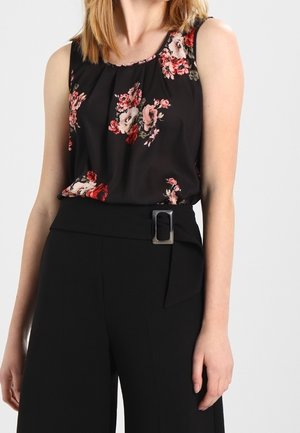 Woman wearing a sleeveless black top with red and pink floral prints, paired with high-waisted black pants featuring a wide belt with a silver buckle.
