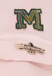 Rosa stofkasket med en broderet grøn og gylden "M" på fronten og et beige "AIRFORCE" vinge-logo på siden.