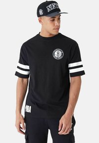 Schwarzes T-Shirt mit kurzen Ärmeln und weißen Streifen an den Ärmeln, kreisförmiges "Brooklyn New York"-Grafikdesign auf der Brust und "NBA"-Text an der Seite.