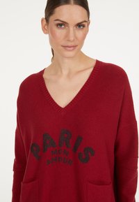 Roter Pullover mit V-Ausschnitt und lockerer Passform, mit einem grafischen Druck "PARIS MON AMOUR" in Schwarz und zwei vorderen Taschen. Glatte Textur.