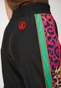 Schwarze Sportshorts aus strukturiertem Material. Mit einem mehrfarbigen Leopardenmuster an der Seitennaht, grünen und orangefarbenen Streifen sowie einem Logo-Patch.