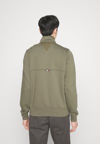 Oliwkowa bluza z wysokim kołnierzem, z trójkątnym karczkiem, ściągaczami przy mankietach i małym wielokolorowym logo blisko tylnego szwu.