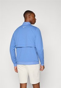 Giacca a pullover blu Nike con colletto alto, maniche lunghe e logo bianco sulla schiena. Presenta un tessuto testurizzato e una vestibilità rilassata.