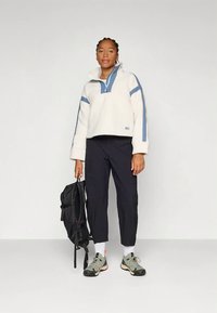 Pulover iz flisa v kremni barvi z modrimi poudarki, z vodilnim ovratnikom z zadrgo na polovici; kombinirano s črnimi hlačami. Črni nahrbtnik in sivi supergi z večbarvnimi podplati.