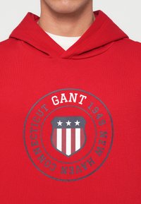 Sweatshirt vermelho feito de algodão, com um gráfico circular que apresenta um escudo, três estrelas e o texto "GANT 1949 NEW HAVEN CONNECTICUT."