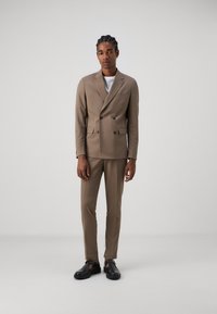 Abito beige doppiopetto con vestibilità su misura. La giacca presenta una tasca, mentre i pantaloni sono a taglio slim. Completato con scarpe nere.