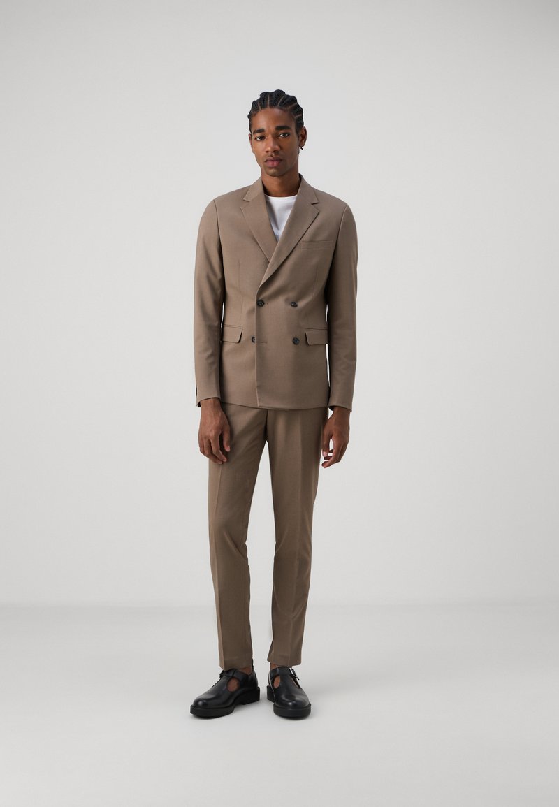 Abito beige doppiopetto con vestibilità su misura. La giacca presenta una tasca, mentre i pantaloni sono a taglio slim. Completato con scarpe nere.